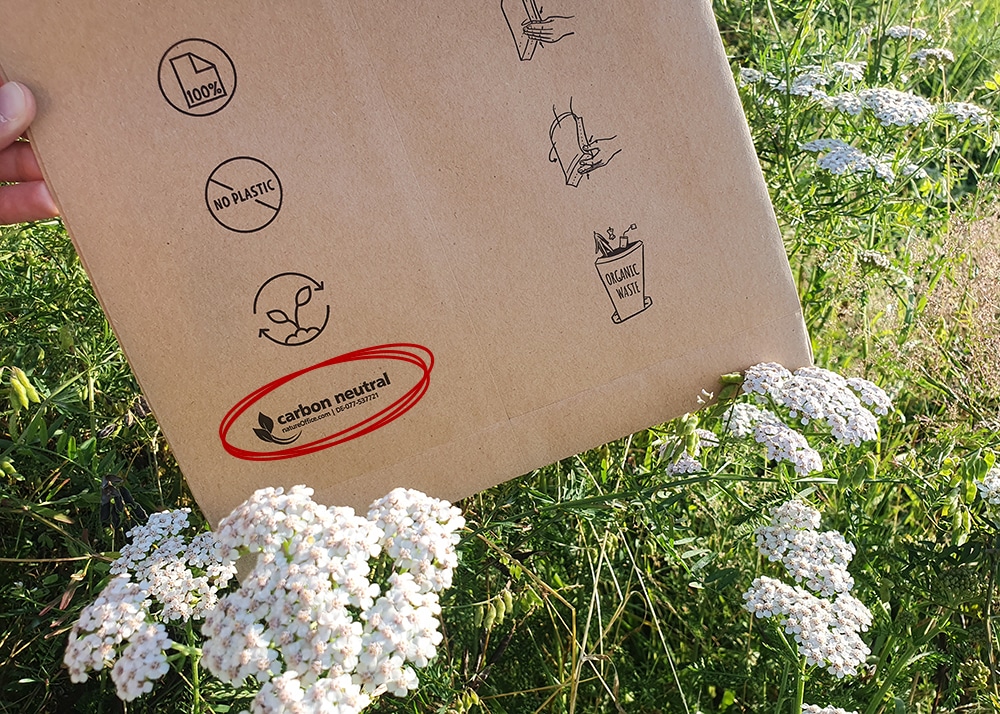 A brown paper mailing bag (DEBAPOST Paper Second Life) with sustainability logos and labelling is pictured in a meadow, surrounded by white flowers. The logos include “100% paper”, “no plastic", “organic waste”. In addition, there is a “carbon neutral” logo, circled in red.